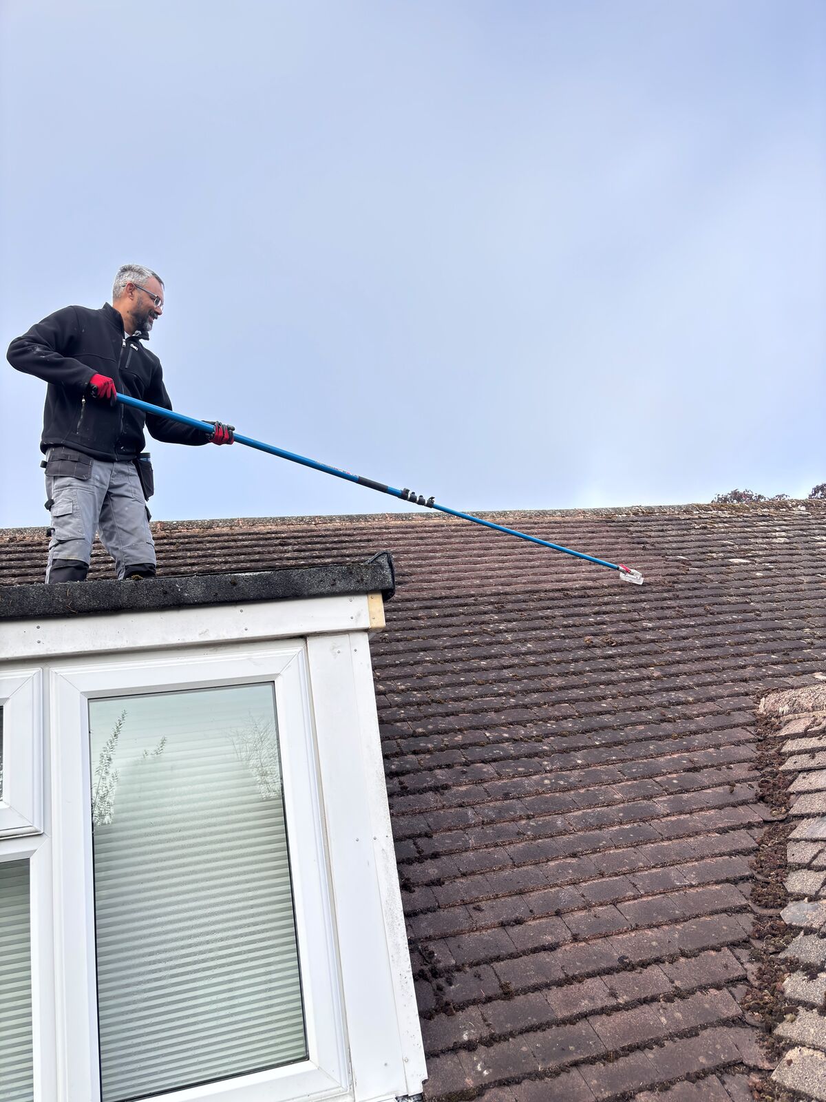 Roof cleaner hand-scraping moss from concrete roof tiles with a long-handled scraper